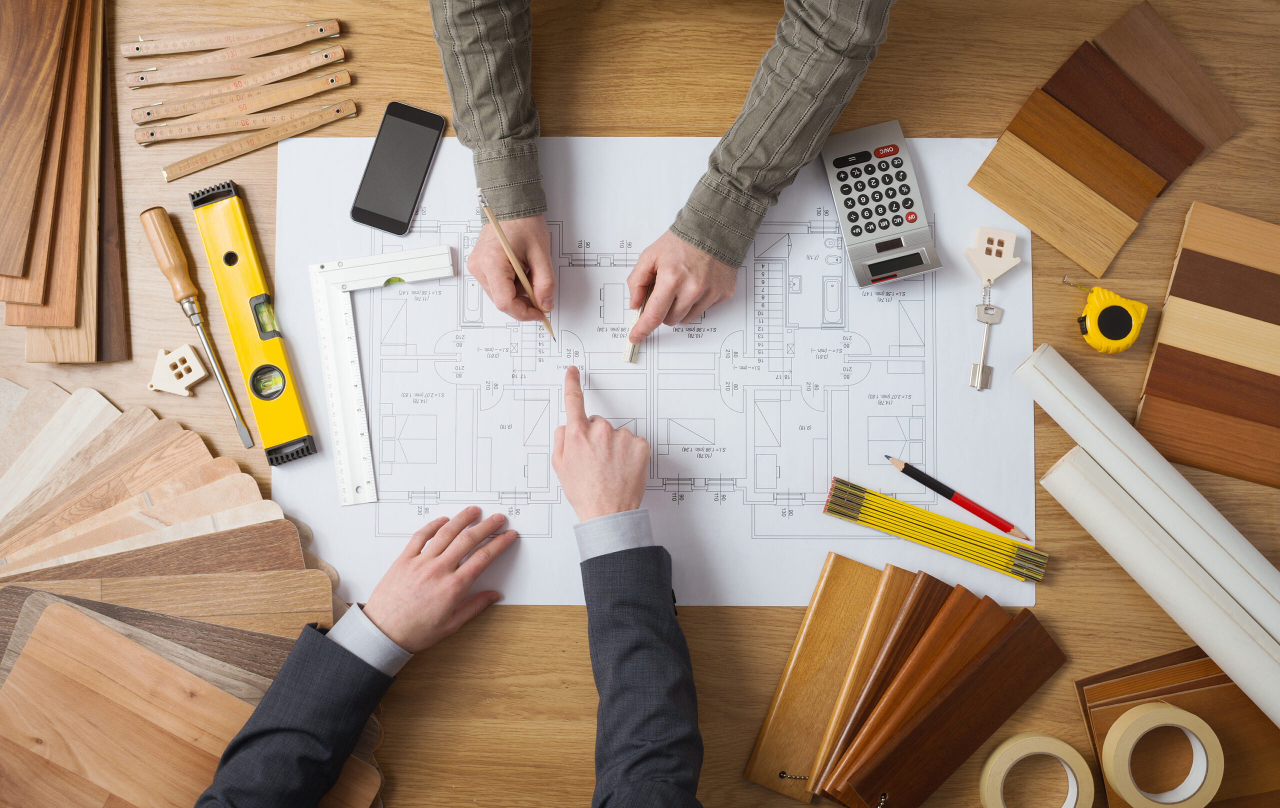 Auf einem Tisch liegen eine Bauzeichnung, ein Taschenrechner, Werkzeuge und Holzproben. Zwei Männer, von denen nur die Arme zu sehen sind, arbeiten an dem Plan. Das Bild symbolisiert die Tätigkeit von Architekten.